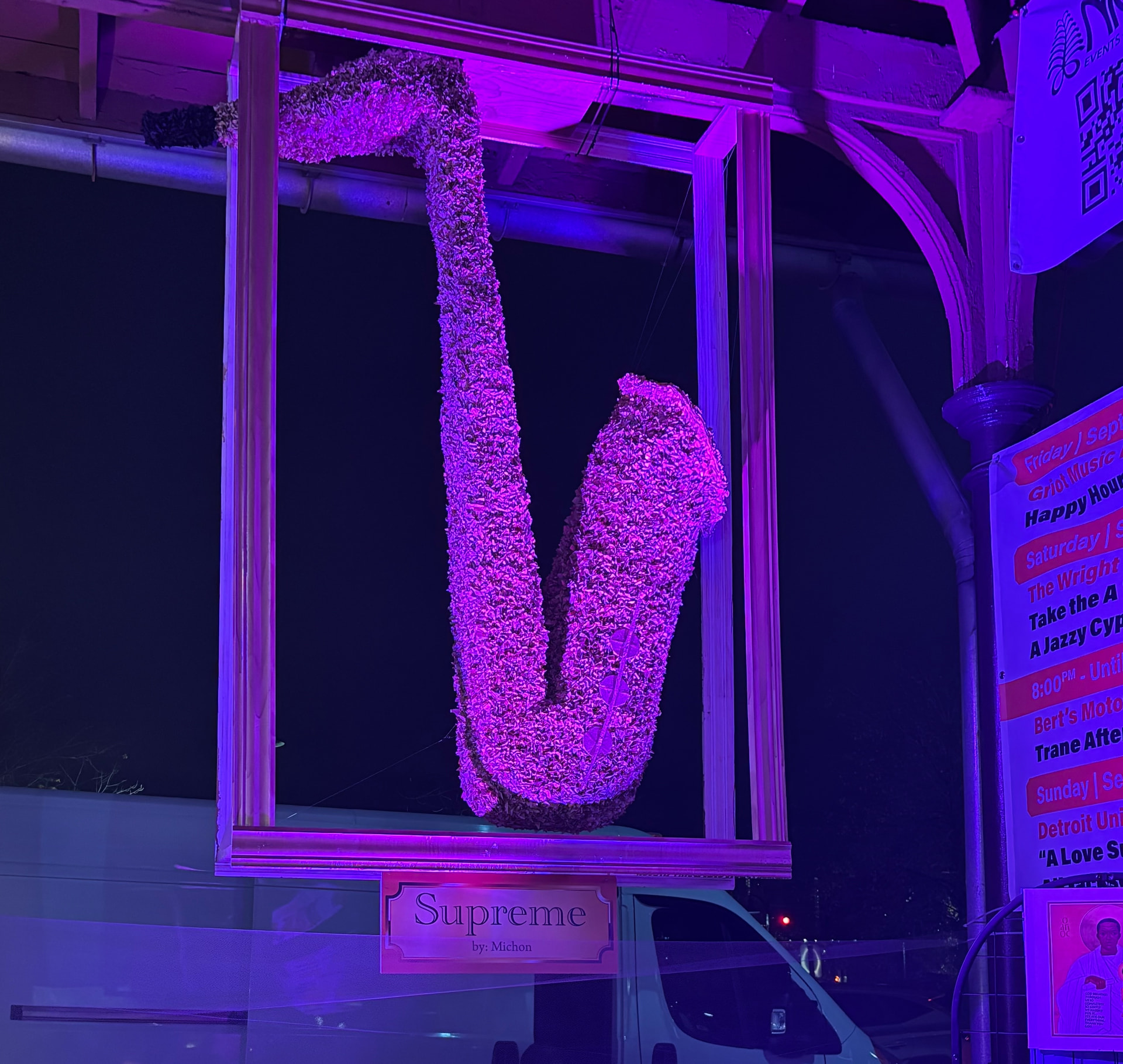 Large handmade tissue-paper saxophone sculpture titled 'Supreme' suspended in a gold frame, lit in purple at an evening event